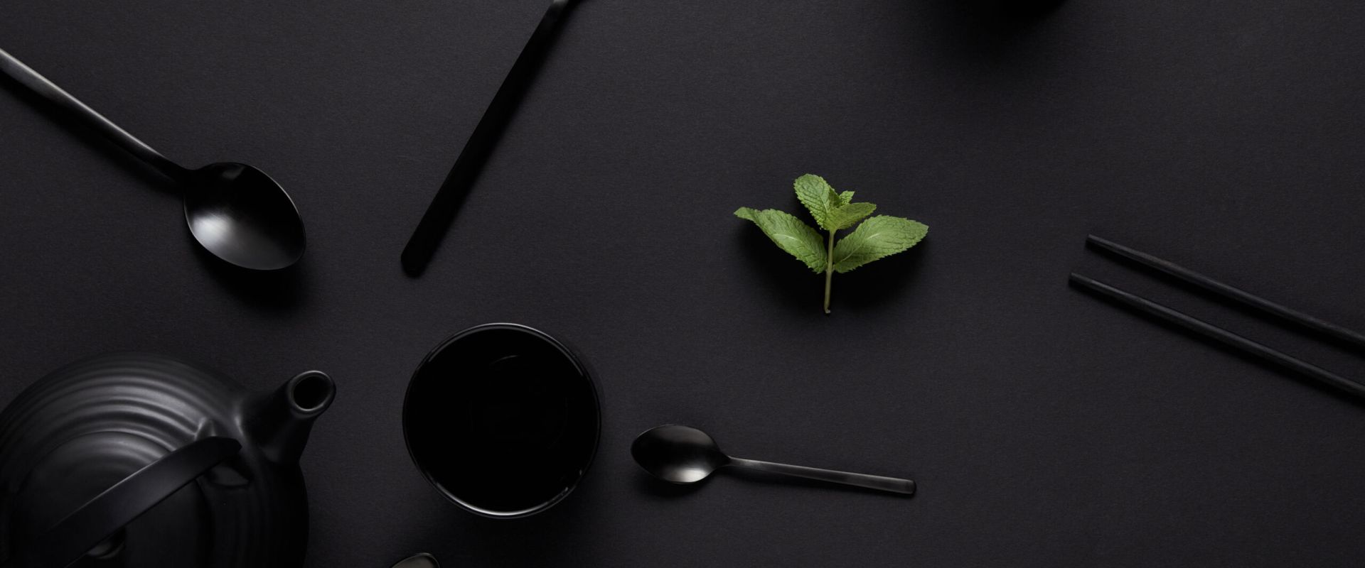 Top view of black teapot, chopsticks, various spoons, fork, cup and mint leaves on black table
